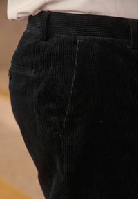 Mustat corduroy-housut, joissa on pystysuorat raidoitukset, tasainen etuosa ja sivutasku napilla. Vyötärönauha on teksturoitu.