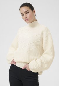 Cream knitted sweater with high ribbed collar, textured diamond pattern, and oversized sleeves; paired with black pants.
