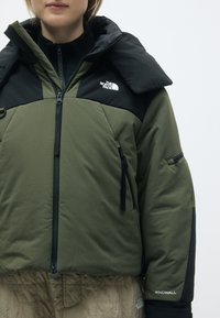 Casaco verde e preto com um grande colarinho, fecho com zip, e múltiplos bolsos, feito de um tecido texturizado, apresentando um logotipo da marca.