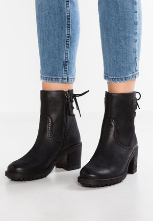 Stivali neri in pelle fino al polpaccio con tacchi a blocco, cerniere laterali e dettagli in pizzo sul retro, indossati con jeans in denim blu a vita corta su un pavimento bianco.