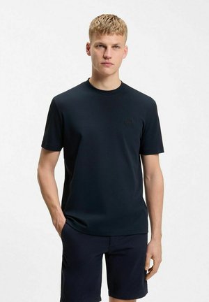 Jonge man met kort blond haar die een effen donkerblauw T-shirt met korte mouwen en bijpassende korte broek draagt, staat met één hand in zijn zak.