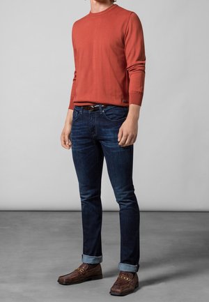 Man met een roestoranje overhemd met lange mouwen, donkerblauwe opgerolde spijkerbroek, bruine leren riem en bruine leren loafers, staand op een grijze vloer.