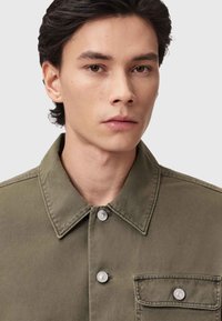 Camicia verde oliva con colletto a punta, tasca frontale e dettagli in metallo sui bottoni, realizzata in un tessuto durevole con una consistenza liscia.