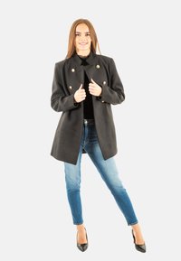 Abrigo negro de doble botonadura con botones dorados, que presenta un cuello alto y una tela suave. Combinado con jeans skinny azules y tacones negros.