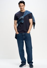 T-shirt navy, jeans blu scuro dal taglio comodo e sneakers sportive, con una giacca di jeans appoggiata su una spalla. Texture lisce in tutto.