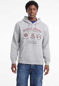Felpa grigia a strati con il logo "Guess Jeans" e i simboli in rosso e blu navy. Il tessuto sembra morbido e presenta una tasca frontale.