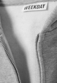 Sweat à capuche gris zippé avec une texture de tissu lisse, une fermeture éclair en métal et une étiquette "WEEKDAY" à l'intérieur du col.