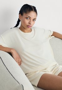 Beige bordázott kötött felső, laza szabással és rövid ujjal, párosítva az azonos színű rövidnadrággal. A modell keze egy textúrált világosszürke kanapén nyugszik.