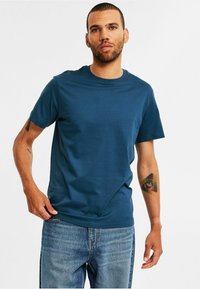 Mand med kort hår iført en blå T-shirt og jeans, der viser en farverig trekantet øjetatovering på sin højre underarm.