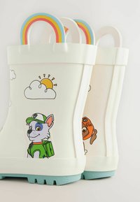 Bottes de pluie en caoutchouc blanches avec des poignées arc-en-ciel, ornées de dessins de chiens de dessin animé et de graphismes de soleil/nuages. Semelles texturées de couleur bleu clair pour une meilleure adhérence.