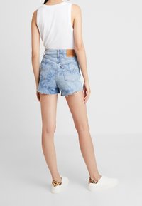 Shorts de mezclilla en azul claro con dobladillo deshilachado, que presentan un patrón sutil y una etiqueta de cuero en la parte trasera. Combinados con zapatillas blancas con acentos de leopardo.