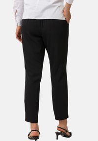 Svarta, smala byxor med en slät yta och elastisk midja, tillsammans med svarta, remmade klacksandaler. Bakvy, tydlig silhuett.