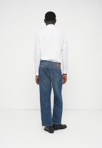 Εφαρμοστή, λευκή μπλούζα με μακρύ μανίκι συνδυασμένη με μπλε τζιν παντελόνι που διαθέτει δερμάτινο λογότυπο στη μέση πίσω και δύο πίσω τσέπες.