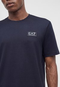 Άνδρας που φοράει σκούρο μπλε μπλουζάκι με μικρό λογότυπο EA7 Emporio Armani στην αριστερή πλευρά του στήθους.