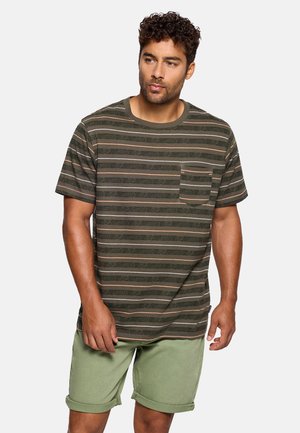 Hombre con cabello rizado que lleva una camiseta de manga corta a rayas verdes y marrones con bolsillo en el pecho y pantalones cortos verde claro remangados.