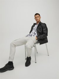 Black leather jacket over a white-and-blue plaid shirt, cream pants with elastic cuffs, and black lace-up sneakers, seated on a metal chair.