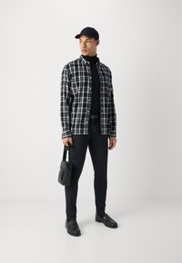 Camicia a quadri bianco e nero sopra un dolcevita nero, pantaloni neri aderenti, scarpe nere e una piccola borsa nera. La modella è in piedi contro uno sfondo bianco.