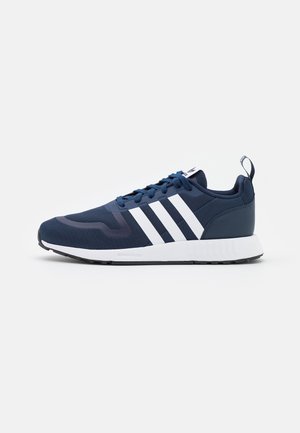 Baskets de sport bleu marine avec des bandes blanches, lacets, col rembourré et semelle blanche sur un fond clair uni.