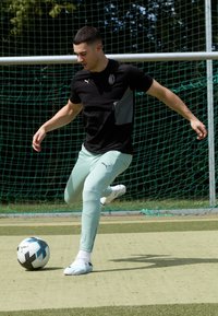 Camisa atlética negra y gris, pantalones verde menta y zapatos de fútbol blancos. Pateando un balón de fútbol negro y blanco en un campo de césped artificial.