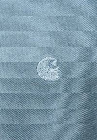 Modra tkanina z teksturirano strukturo, s belim vezenim logotipom, ki spominja na spiralno školjko v spodnjem levem kotu.