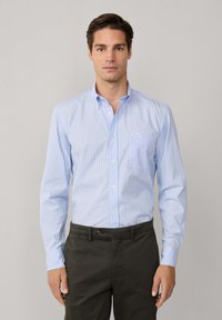 Camicia azzurra a righe con taschino, abbinata a pantaloni verde scuro. Presenta un colletto a punta e bottoni bianchi.