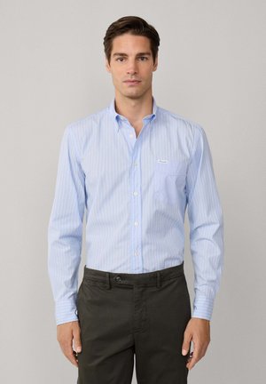 Camisa azul claro a rayas con un bolsillo, combinada con pantalones oscuros verdes. Presenta un cuello extendido y botones blancos.