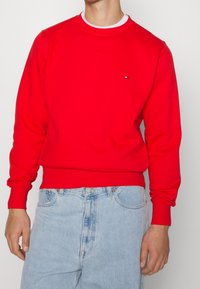 Rode sweatshirt met geribbelde manchetten en onderkant, ronde halslijn en klein logo op de borst. Gecombineerd met lichtblauwe spijkerbroek.