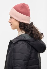 Gorro de punto con parte superior en burdeos y banda rosa, forma redondeada, material texturizado y acabado suave. Lleva puesto con una chaqueta de plumas negra.