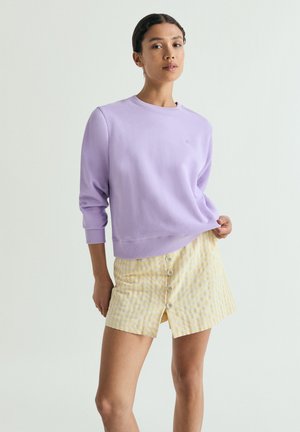 Femeie purtând un sweatshirt lavandă cu mâneci lungi și o fustă galbenă în carouri, cu nasturi în față, stând pe un fundal simplu și deschis la culoare.