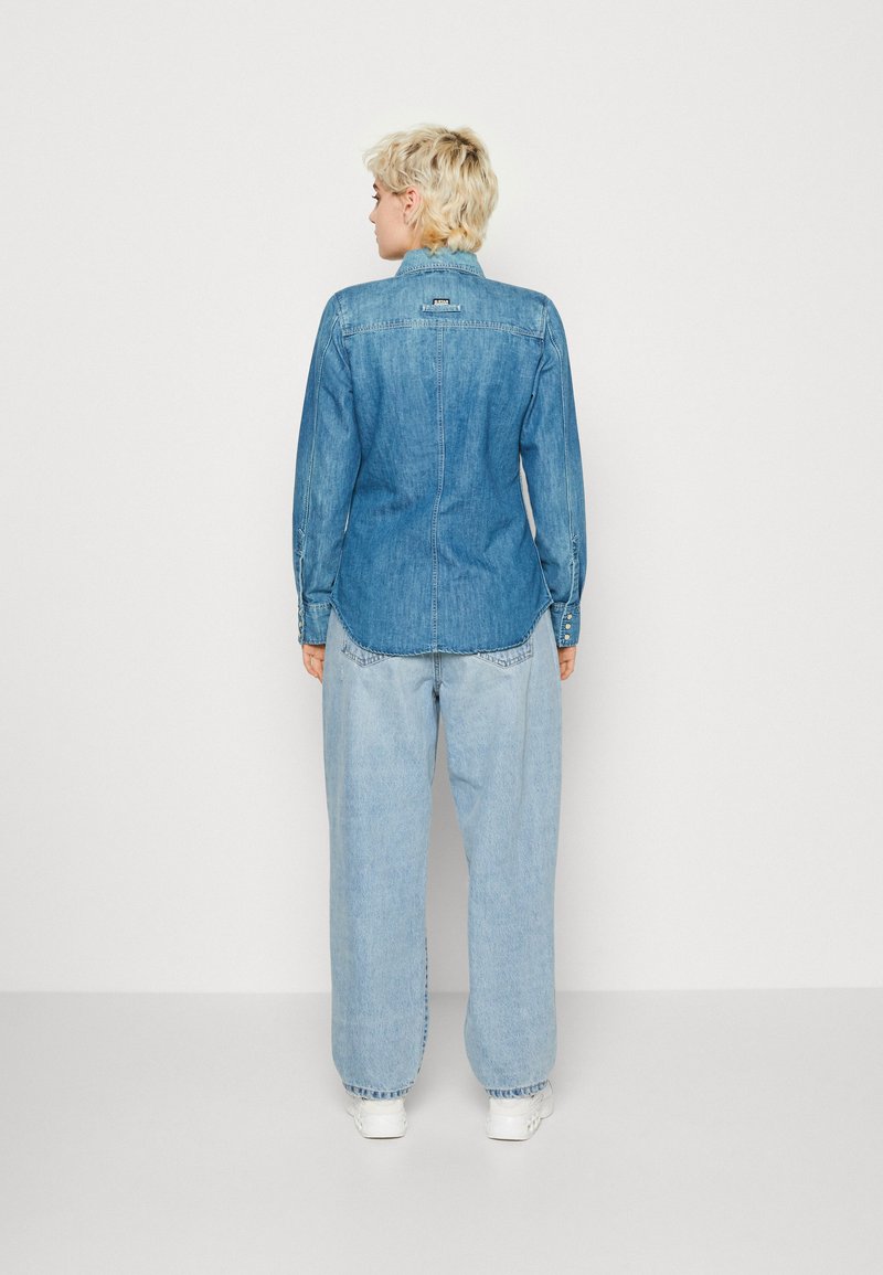 Helles blaues Denim-Hemd mit Kragen, tailliertem Schnitt und Knopfdetails an den Manschetten, getragen mit lockeren, hellblauen Jeans.