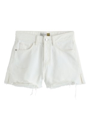 Kvinde iført en ærmeløs strikket top og hvide shorts, der holder en stor vævet strandtaske med skaldyrsdekorationer mod en klar himmel baggrund.