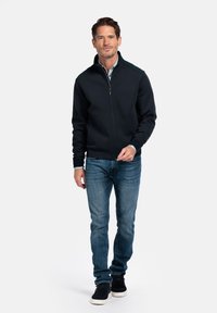Heren donkerblauwe ritsjack met een hoge kraag, geribde manchetten en contrasterende schouderpanelen, gecombineerd met lichtblauwe jeans en zwarte schoenen.