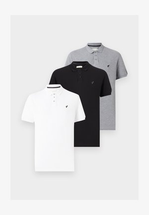 Homme debout, les mains dans les poches, portant un polo blanc, un jean bleu clair et des baskets noires sur un fond blanc.