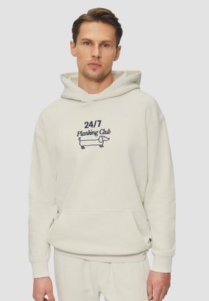 Crèmekleurige hoodie met een kangoeroezak, met geborduurde tekst "24/7 Planking Club" en een lijntekening van een hond. Zachte textuur en een ontspannen pasvorm.