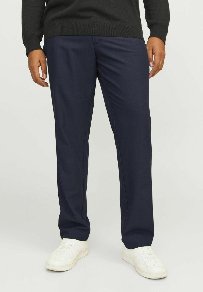 Marineblaue Hose aus glattem Material, taillierte Passform, mit geradem Bein-Design und dezenten Faltendetails. Getragen mit weißen Sneakers.