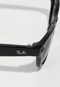 Occhiali da sole in plastica nera con una montatura spessa, dotati di un accento in metallo argentato e del logo "Ray-Ban" inciso sul braccio. Include lenti scure.
