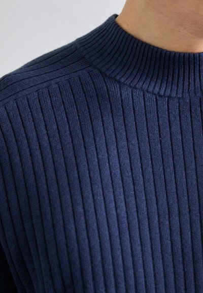 Marineblå ribbestrikket genser med høy krage. Har vertikale mønstre, myk tekstur og en tettsittende design som former seg etter kroppen.