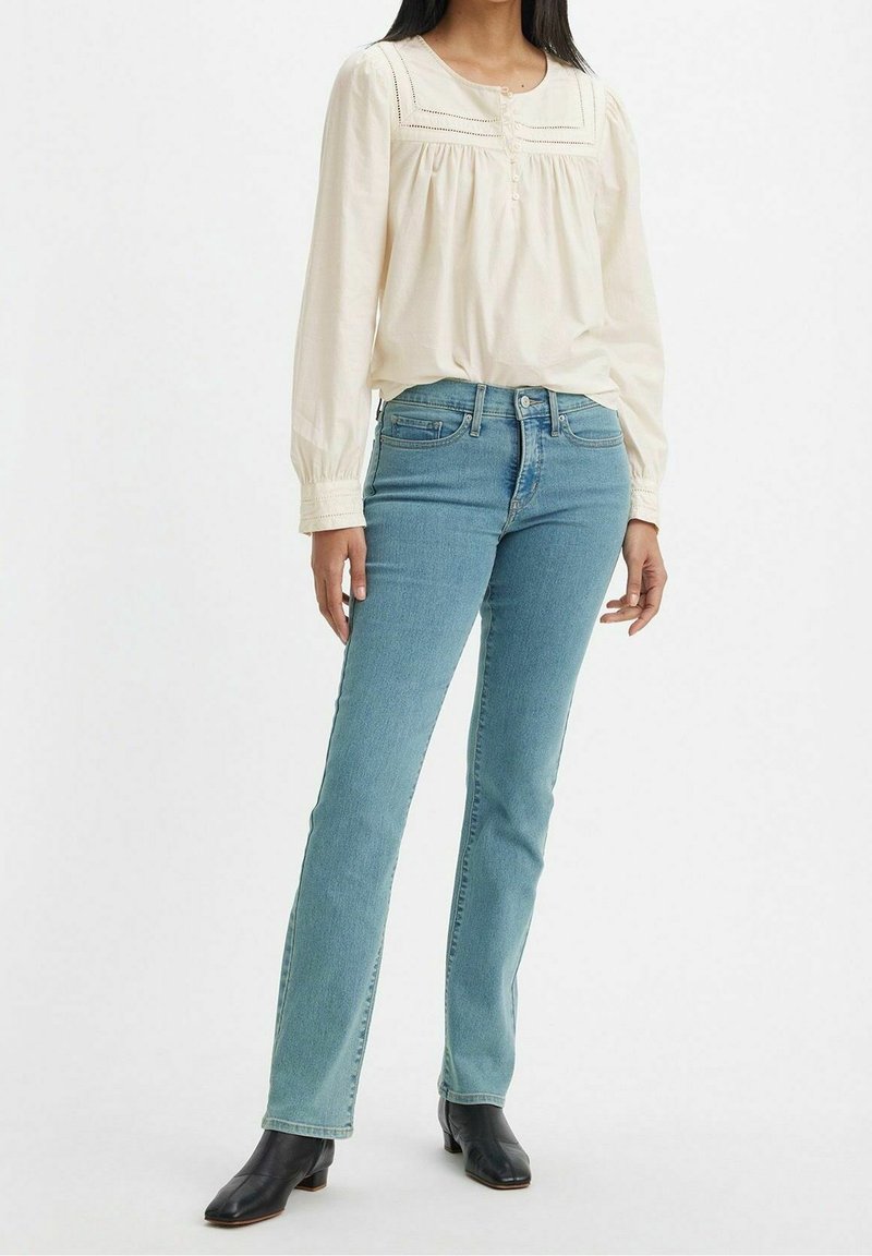 Blouse légère à manches longues de couleur crème avec des détails froncés et des boutons, associée à un jean droit bleu clair. Bottines noires à bout pointu.