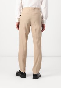 Pantalon beige ajusté avec un devant plat, deux poches arrière et une coupe droite, associé à des chaussures noires. Texture de tissu lisse.