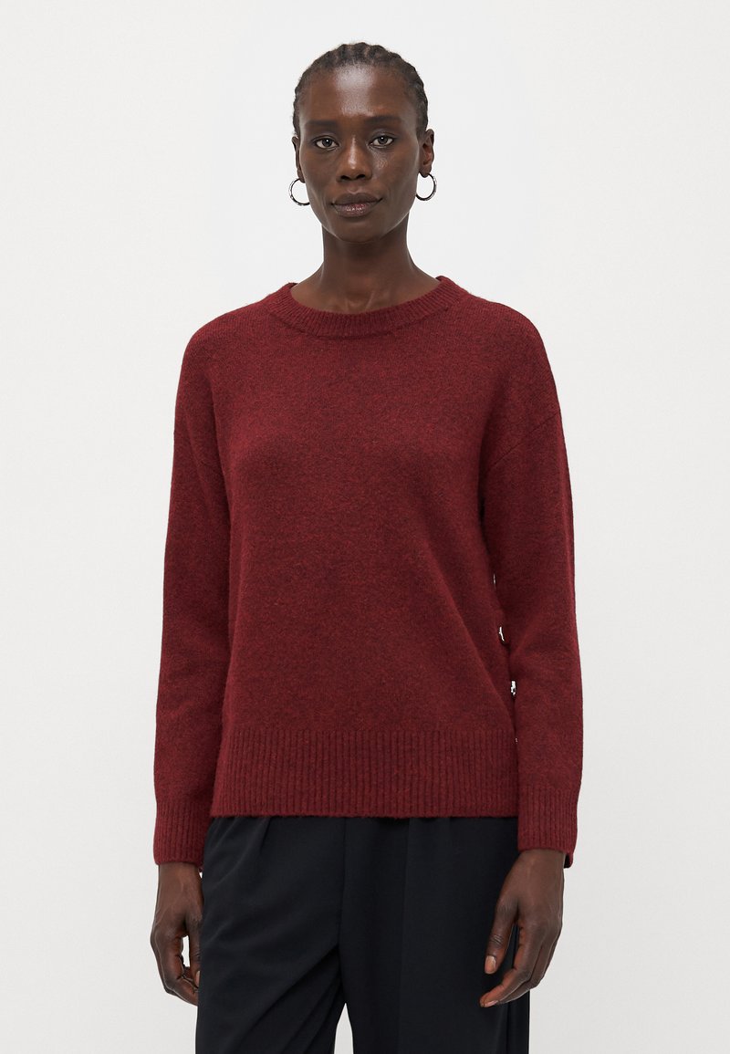 Maglione lavorato a maglia bordeaux con scollatura tonda, orlo e polsini a costine, texture morbida e design con spalla scesa. Indossato sopra pantaloni scuri.
