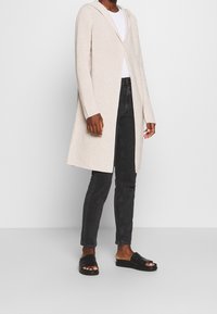 Long gilet en tricot beige avec capuche, à l'avant ouvert, texture côtelée. Associé à un jean skinny noir et des sandales noires sans lacets.