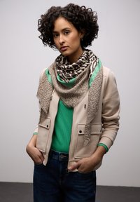 Femme aux cheveux bouclés portant une veste beige, un pull vert, une écharpe à motif animalier avec une bordure verte, et un jean foncé, les mains dans les poches, fond neutre.