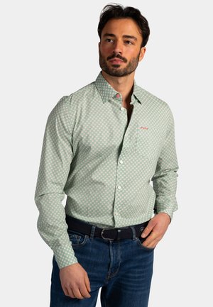 Man met een lichtgroen overhemd met patroon en borstzak, een donkere geweven riem en een blauwe spijkerbroek, staand met één hand in de zak.