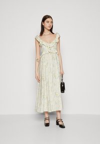 Robe maxi florale en crème douce, avec un col et une taille volantés, mettant en avant des motifs floraux jaunes et bleus. Assortie à des talons noirs.