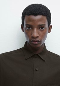 Brown button-up shirt with a pointed collar, featuring a smooth texture and a single chest pocket. Simple design with understated details.