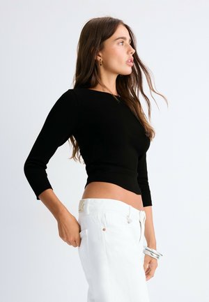 Žena s dlhými hnedými vlasmi, ktorá má na sebe čierny crop top a biele nohavice, pozerá sa do strany s jednou rukou v kapse.