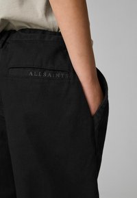 Črne hlače prikazane s bližnjim pogledom na zadnji žep, ki ima všit napis "ALLSAINTS". Tkanina je gladka in mat.