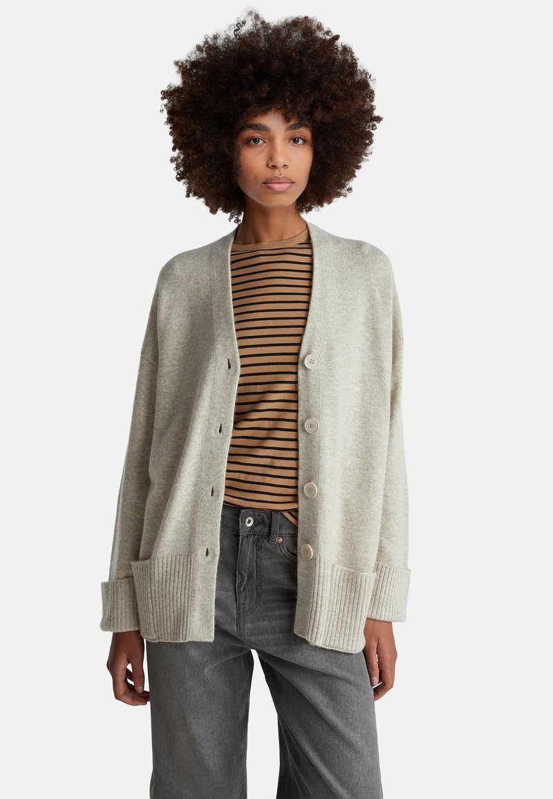 Cárdigan de punto gris claro con puños y dobladillo acanalados, con botones en la parte delantera. Llevado sobre una camiseta de rayas marrón y beige con un corte relajado.