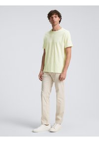 Lys gul bomull T-skjorte med korte ermer og rund hals, kombinert med beige bukser og hvite joggesko. Enkel design, ingen mønstre.