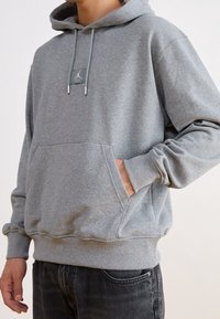 Grå huvtröja i mjukt tyg, med en känguruficka, dragsko i huvan och en liten logotextil på bröstet. Ribbstickade mudd och nederkant.